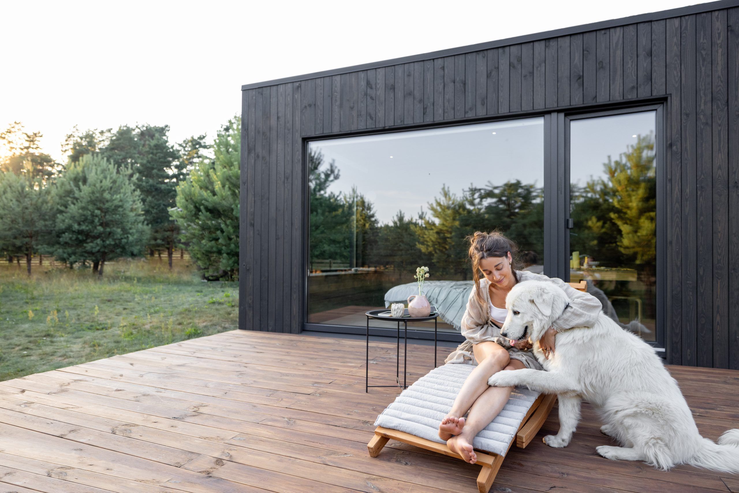Woman relaxing on a cushioned wooden deck chair outside a modern black timber garden studio with a large sliding glass window. She is smiling and gently interacting with a large white dog sitting beside her with its paw on her leg.