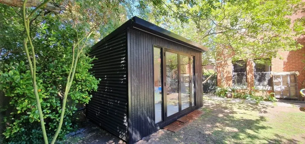 Small modern black garden studio shed with vertical cladding and glass double doors, set in a leafy backyard with trees, grass, and a brick house in the background.